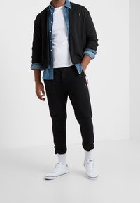 Man i lager-på-lager-casual outfit med svart zip-up jacka, denimskjorta, vit t-shirt, svarta joggers, vita strumpor och vita sneakers.