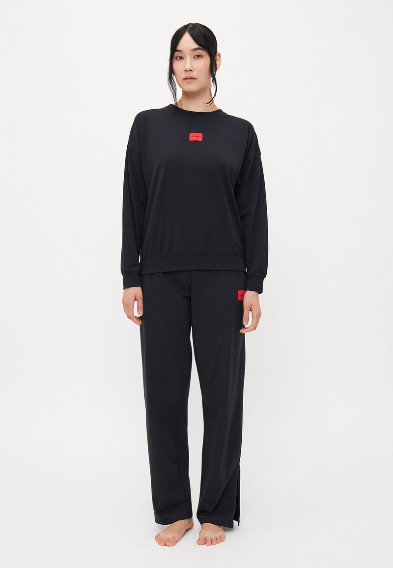 Zwart sweatshirt en broekset gemaakt van gladde stof. Het sweatshirt heeft een rood logo-patch; de broek heeft zij-splitten en een bijpassend logo.