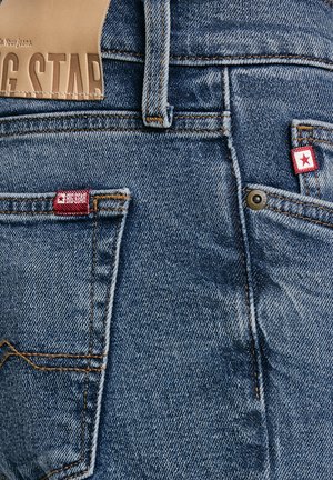 Sinine teksapükste tagataskute lähivaade, millel on punase tähega kaubamärgiga sildid ja kontrastset oranži õmblust.