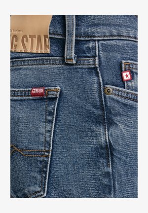 Zbliżenie na tylne kieszenie niebieskich dżinsów z metkami z czerwoną gwiazdą oraz kontrastowymi pomarańczowymi przeszyciami.