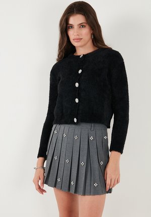Black, fluffy cropped cardigan with silver flower-shaped buttons, paired with a gray pleated skirt adorned with floral embellishments.