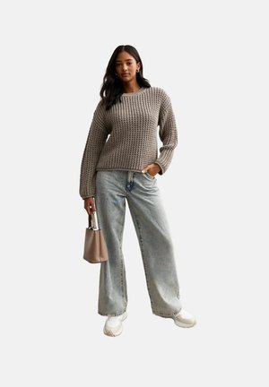Beżowy sweter dzianinowy z teksturowanym wzorem, zestawiony z jasnoniebieskimi spodniami typu wide-leg i białymi sneakerami. Modelka trzyma małą beżową torebkę.
