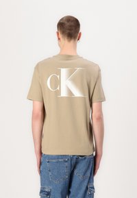 Šviesiai ruda medvilninė marškinė su dideliu baltu "cK" logotipu ant nugaros. Trumpomis rankovėmis ir klasikiniu siluetu. Dėvimi su mėlynomis kargo stiliaus džinsais.