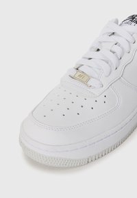 Primo piano di una sneaker Nike Air Force 1 bianca che mostra la punta perforata, i lacci bianchi con il tag metallico "AF1" e il bordo della suola testurizzato.