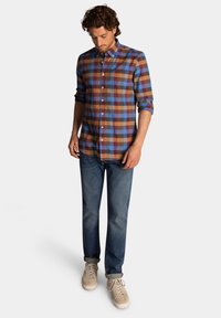 Geruite shirt in blauwe, bruine en rode tinten; lange mouwen, knoopsluiting aan de voorkant, één borstzak, gecombineerd met blauwe jeans en beige sneakers.