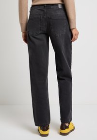 Persona vestida con jeans rectos negros, suéter beige y zapatillas amarillas, de pie de espaldas sobre un fondo blanco liso.