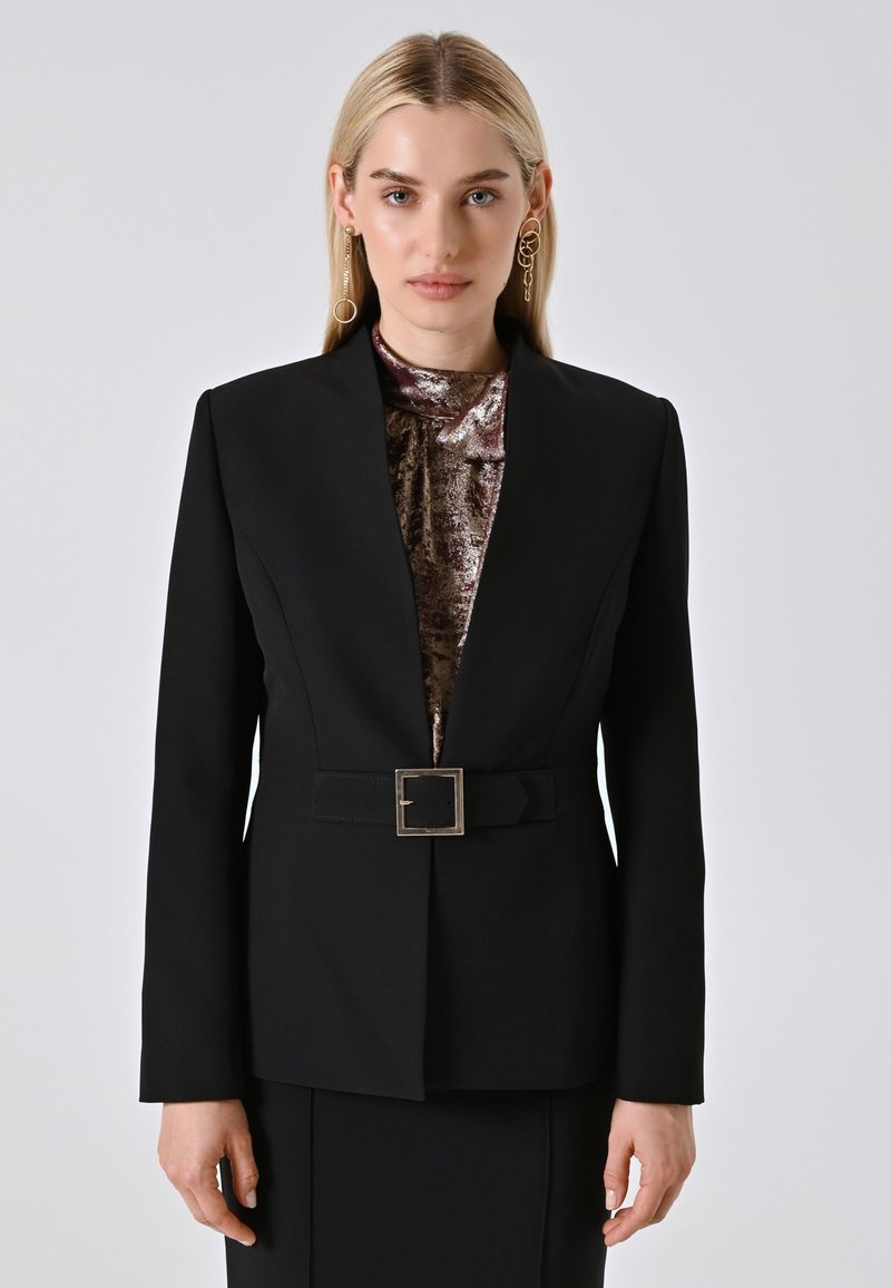 Donna bionda che indossa un blazer nero con cintura sopra un top metallico bronzo, con orecchini a cerchio dorati non abbinati, in piedi contro uno sfondo semplice.