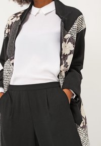 Manteau patchwork noir avec des motifs floraux et pied-de-poule, associé à une chemise blanche à col et un pantalon noir. Le tissu semble texturé.