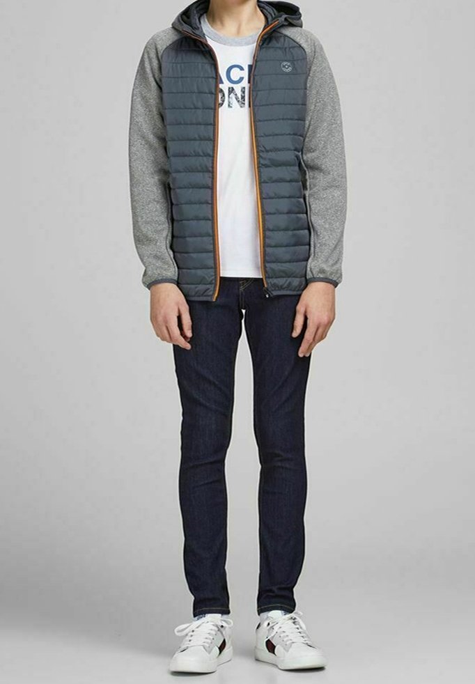 Ocieplana szara kurtka z kapturem, z jednolitą częścią główną i szarymi rękawami, zestawiona z ciemnymi dżinsami i białymi trampkami.