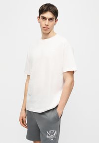 Νεαρός με κοντά καστανά μαλλιά, ντυμένος με λευκό μπλουζάκι και γκρι σορτς New Balance, στέκεται με το ένα χέρι στην τσέπη σε απλό φόντο.