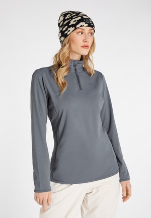 Frau trägt einen grauen Pullover mit langen Ärmeln und Viertelreißverschluss, ein gemustertes schwarz-beiges Beanie und helle Hosen, schaut nach rechts.