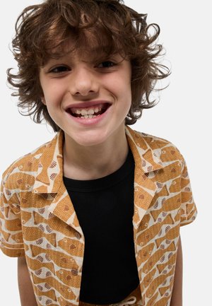 Enfant souriant aux cheveux bruns bouclés, portant une chemise orange à motifs ouverte sur un t-shirt noir, face à un fond uni.
