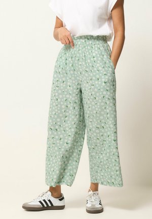 Personne portant un pantalon large vert clair à imprimé marguerites blanches, une chemise blanche à manches courtes et des baskets blanches avec des rayures noires.
