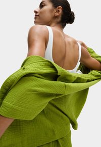 Camicia verde strutturata appoggiata sulla spalla, abbinata a una canotta bianca a coste. La camicia presenta un bottone e un design dal taglio morbido.