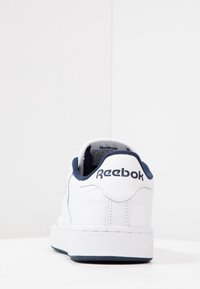Vit sneakers med en slät läderöverdel, marinblå detaljer vid hälen och en strukturerad gummisula. Har Reebok-loggan på baksidan.