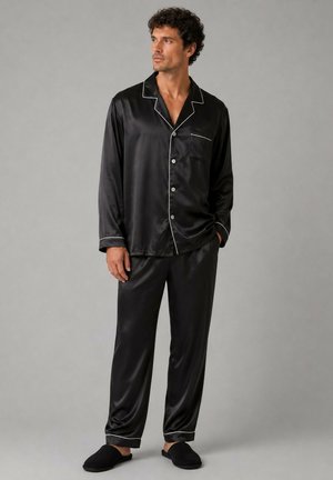 Homme portant un pyjama noir en satin à manches longues boutonné avec un passepoil blanc, pantalon assorti et chaussons noirs, debout devant un fond gris uni.