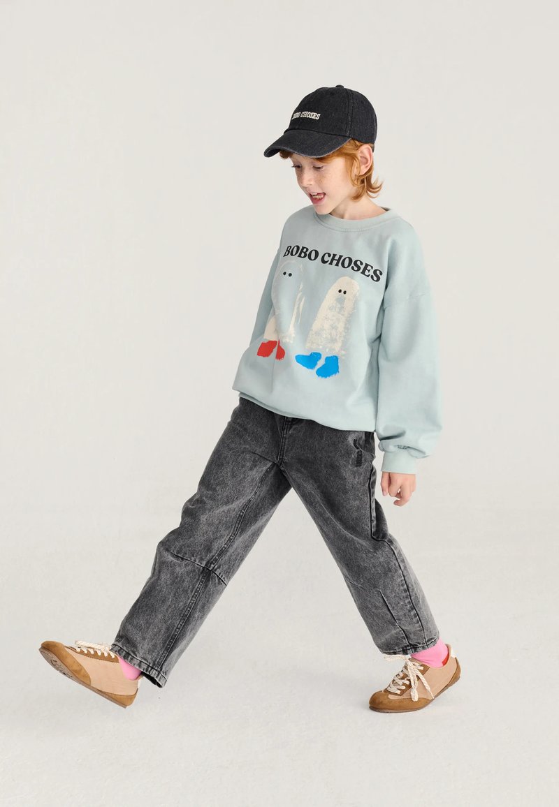 Bambino che indossa una felpa azzurra con stampe di fantasmi, jeans grigio scuro, scarpe marroni, calzini rosa e un berretto nero, cammina con una gamba sollevata.