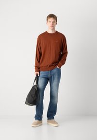 Maglione a maglia marrone, jeans blu, sneaker beige. Il modello tiene una borsa nera. Fit casual, maniche lunghe, polsini a costine. Design semplice e pulito.