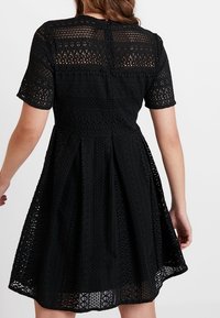 Schwarzes Spitzenkleid mit kurzen Ärmeln, das ein filigranes Laser-Cut-Muster aufweist, einen taillierten Oberkörper und einen ausgestellten Rock hat. Rückenschluss am Halsauschnitt.
