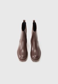 Une paire de bottes chelsea en cuir marron avec des tirants et des panneaux élastiques sur un fond blanc uni.