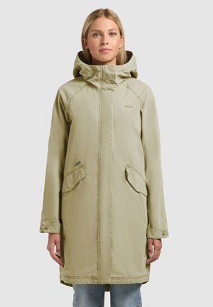 Kobieta ubrana w długi, jasnobeżowy płaszcz z kapturem, z klapkowymi kieszeniami i mankietami na zatrzaski, stojąca na tle jednolitego jasnego tła.