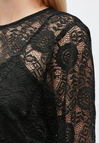 Juoda nėriniuota palaidinė su gėlių ir geometriniais raštais, permatoma tekstūra, apvaliu kaklu ir ilgomis rankovėmis.