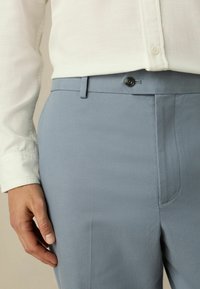 Lichtblauwe broeken van een gladde stof, met een knoop en een voorkeep. De tailleband is plat met een subtiele textuur.