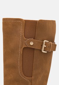 Bota de ante marrón con una pantorrilla ancha, que presenta un panel de tela y un detalle de hebilla dorada. La costura en contraste añade textura.