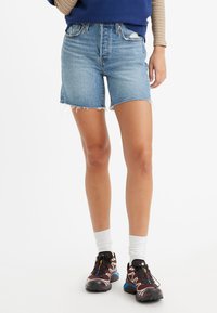 Shorts de mezclilla en azul claro con dobladillos deshilachados, presentan una cintura alta y un diseño clásico de cinco bolsillos, combinados con zapatillas deportivas negras y calcetines blancos.