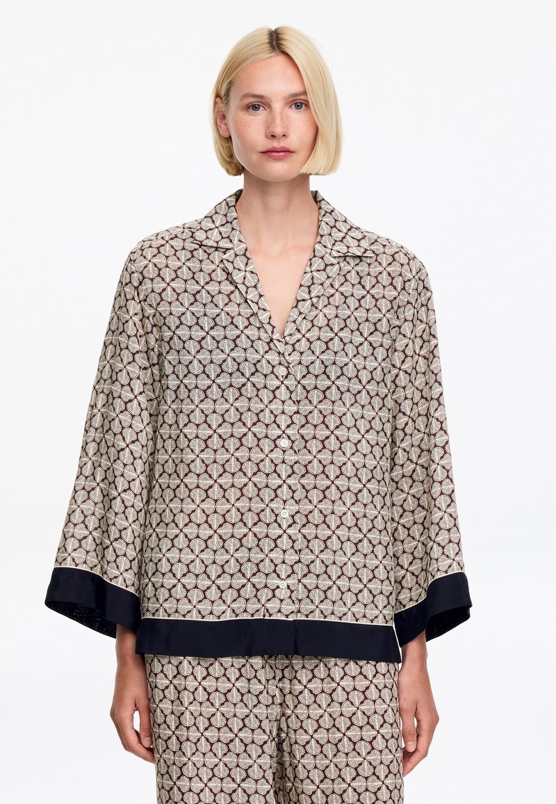 Femme blonde portant un haut boutonné ample à motifs assorti et un pantalon avec un bord noir aux manches et à l'ourlet, debout devant un fond blanc.