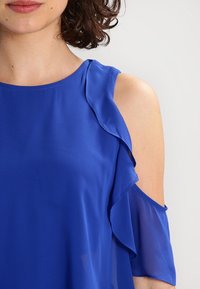 Top azul de hombros al descubierto, hecho de una tela ligera. Presenta acentos con volantes en las mangas, textura suave y un escote redondo.