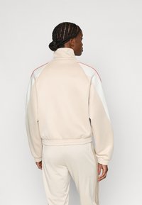 Ανοιχτό μπεζ αθλητικό σακάκι με ψηλό γιακά, ριμπ μανσέτες και λευκές λεπτομέρειες με κόκκινες γραμμές στους ώμους, κατασκευασμένο από λεία ύφη.