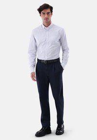 Weißes Hemd mit hellblauen Streifen, kombiniert mit marineblauen Hosen und schwarzen Schuhen. Das Hemd hat einen Kragen und eine Brusttasche.