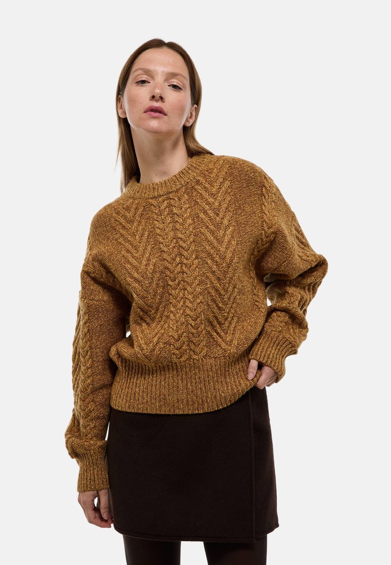 Pull en maille torsadée moutarde avec un col rond et des poignets côtelés, associé à une jupe marron foncé. Texture douce, coupe ample et motif détaillé.