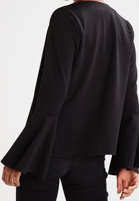 Svart långärmad topp med utsvängda ärmar, tillverkad av mjukt tyg. Baksidan är slät och halsringningen är rundad. Matchas med svarta byxor.