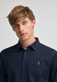 Mörkblå denimskjorta med knäppning, beige sömmar och en liten gul logotyp på vänster bröst. Slät tygtextur.