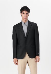 Blazer negro ajustado con dos botones, bolsillos laterales y un sutil detalle de forro, combinado con una camisa gris de botones y pantalones beige.