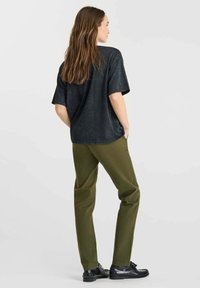 Mujer con cabello largo castaño de pie de espaldas, vistiendo una camiseta negra holgada, pantalones verdes oliva y zapatos negros, contra un fondo sencillo.