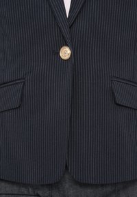 Blazer marine avec des rayures blanches, détails de boutons dorés et deux poches avant. Coupe ajustée avec une texture lisse.