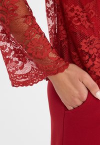 Rode kantmouwen met bloemmotieven, gecombineerd met een effen rode stof eronder, met een zakdetail op het lagere kledingstuk.