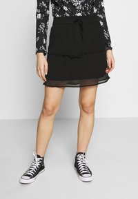 Svart lagerkjol med volangkant, elastisk midja och snörning. Kombinerad med svarta högstövlar med vita sulor.