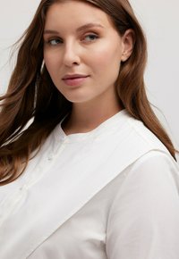 Blusa bianca con bottoni frontali, colletto arrotondato e un ampio dettaglio sulle spalle drappeggiato. Tessuto liscio, design minimale.