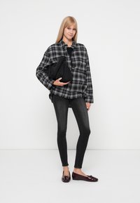 Cămașă oversized cu carouri alb-negru, top negru, blugi skinny negri și pantofi maro lucioși. Model cu o geantă neagră.