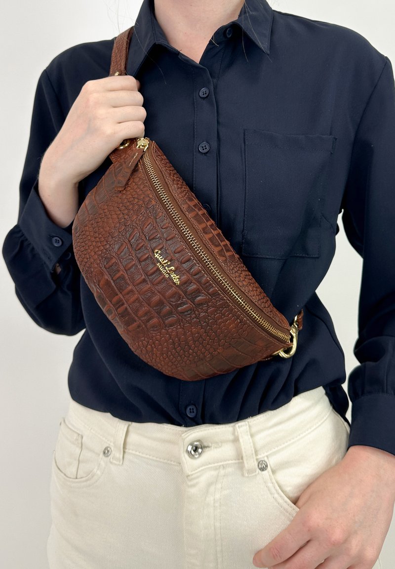 Borsa a tracolla in faux pelle coccodrillo marrone con zip dorata, caratterizzata da una forma arrotondata e una tracolla regolabile, indossata su una camicia blu navy.