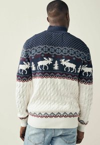 Pull en tricot présentant un haut bleu marine avec un motif de chèvres et d'arbres blancs, une partie inférieure en tricot torsadé blanche, et un col et des poignets côtelés dans des couleurs contrastantes.
