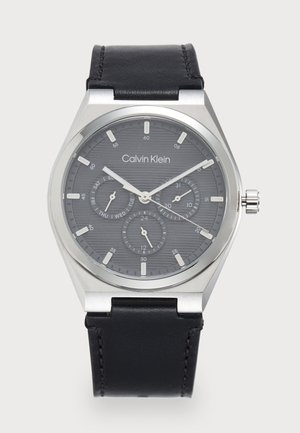 Montre analogique grise avec un cadran texturé, un boîtier en acier inoxydable argenté et un bracelet en cuir noir. Les caractéristiques incluent des sous-cadrans pour les heures, les minutes et les secondes.