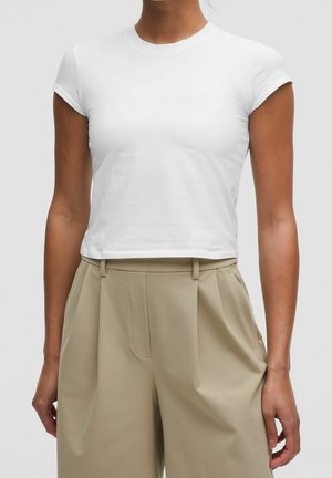 Femme portant un crop top blanc moulant à manches courtes et un pantalon plissé beige taille haute sur un fond uni.