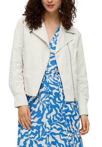 Witte cropped blazer met een notch-kraag, ritsluiting en lange mouwen, gedragen over een blauwe jurk met een wit abstract patroon.