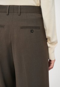Pantalon marron ajusté avec poches passepoilées boutonnées dans le dos, porté avec une chemise crème à manches longues rentrée, montré de la taille à mi-cuisse du côté droit.