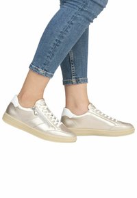 Metallisch goldene Sneakers mit weißen Akzenten, Schnürverschluss und seitlichem Reißverschluss. Glatte Textur und runde Gummisohlen. Getragen mit blauen Jeans.